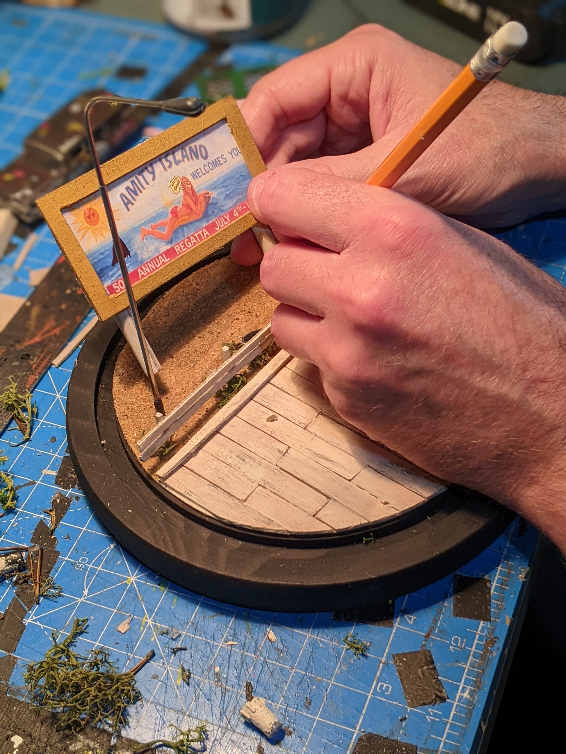 Hand holding a miniature model with a small card labeled 'Amity Island' on a blue cutting mat.